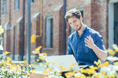 Genç ve yakışıklı bir erkek öğrenci öğretmenleriyle video görüşmesi yaparak bilgisayarla çevrimiçi sınava giriyor. Kampüsün yakınındaki bir bankta oturuyor. Kollarını sallar, açıklar, konuşur.