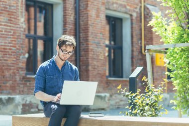 Gözlüklü ve kot pantolonlu genç bir öğrencinin portresi. İnternette bir dizüstü bilgisayarda çalışıyor, ofis merkezinin yanındaki bankta oturmaya odaklanmış..