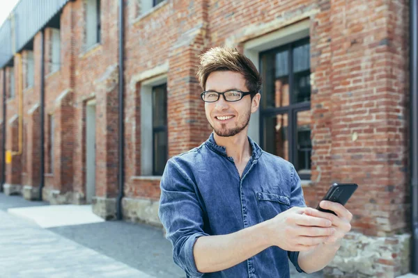 Young handsome young man with glasses travels to another country. Walks around the city and calls on a video call from a mobile phone to friends and family. He waves his hand, greets, shares his impressions