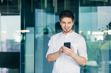 Ofiste pencerenin yanında duran genç işadamı gülümseyen bir cep telefonunun ekranına bakarak online bir mesaj okuyor.