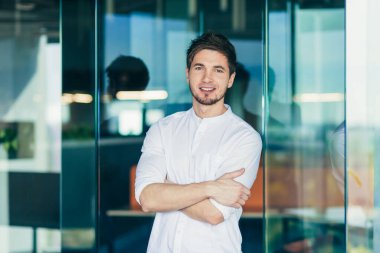 Modern ofisteki genç serbest çalışan, iş adamı portresi. Kameraya kollarını kavuşturup gülümseyen, gömlek giyen bir adam.