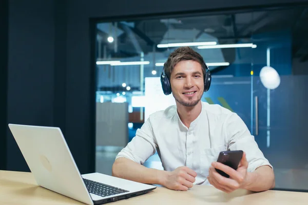 Young businessman works in the office on a laptop, listens to music in big headphones uses the application to listen to online books and podcasts uses the phone
