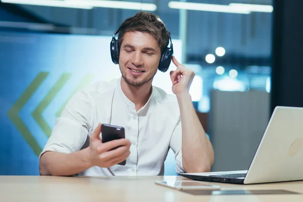 Young businessman works in the office on a laptop, listens to music in big headphones uses the application to listen to online books and podcasts uses the phone