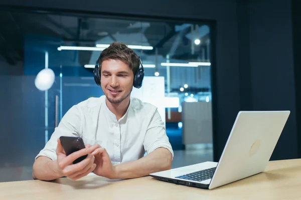 Young businessman works in the office on a laptop, listens to music in big headphones uses the application to listen to online books and podcasts uses the phone