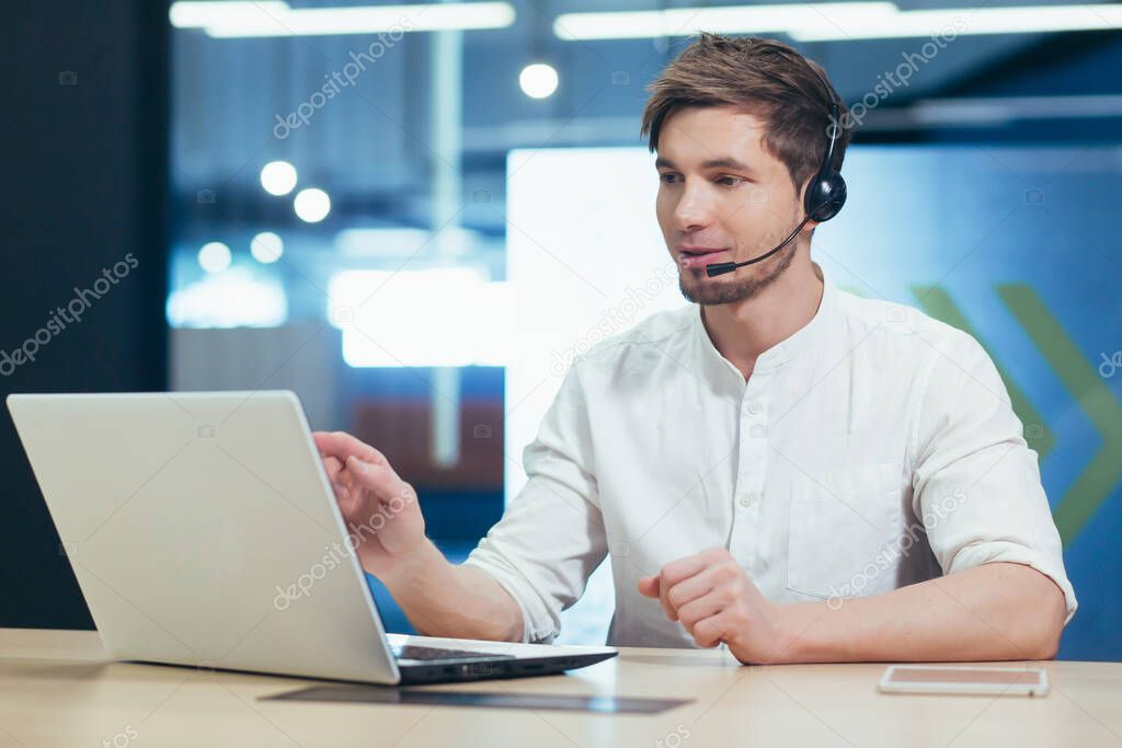 A young office worker working with a laptop uses a headset for video communication, a man from the support service communicates online with the client
