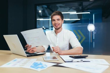 Kağıt işlerinde başarılı bir işadamının portresi, gülümseyen ve elinde tuttuğu belgeye ve mali raporlara bakan bir adam.