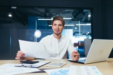 Kağıt işlerinde başarılı bir işadamının portresi, gülümseyen ve elinde tuttuğu belgeye ve mali raporlara bakan bir adam.