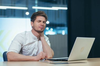 Ofisteki adam yorgun, boynu ağrıyor, gömlekli genç iş adamı sağlıksız bilgisayarla çalışıyor.