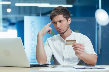 Ofisteki düşünceli ve hayal kırıklığına uğramış genç işadamı online bir mağazadan alışveriş yapmaya çalışıyor, elinde banka kartı olan bir dizüstü bilgisayar kullanıyor.