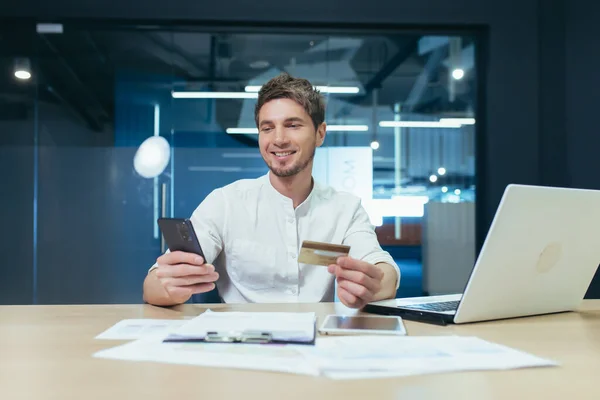 Young man in office working with laptop uses phone and bank credit card for online shopping in online store, happy smiling businessman