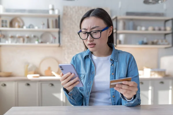 A young Asian woman tries to do a banking operation, uses a phone and a bank credit card, disappointed and unhappy with the result