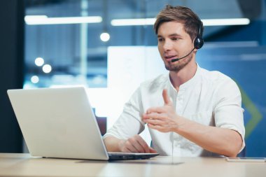 Video konferansı. Kulaklıklı genç yakışıklı bir adam ofiste dizüstü bilgisayarın arkasında oturuyor ve video aracılığıyla iletişim kuruyor..