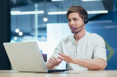 Video konferansı. Kulaklıklı genç yakışıklı bir adam ofiste dizüstü bilgisayarın arkasında oturuyor ve video aracılığıyla iletişim kuruyor. Kameraya bakıyor, gülümsüyor.