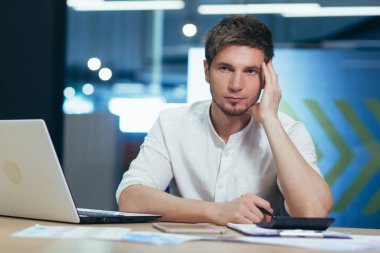 Odaklanmış ve ciddi genç adam bilişim uzmanı, yönetici, iş adamı dizüstü bilgisayar üzerinde çalışıyor ve büroda belgeleri var. Kafasını tutar, düşünür