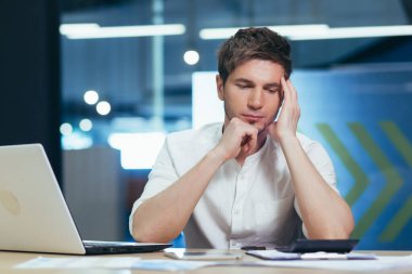 Odaklanmış ve ciddi genç adam bilişim uzmanı, yönetici, iş adamı dizüstü bilgisayar üzerinde çalışıyor ve büroda belgeleri var. Kafasını tutar, düşünür