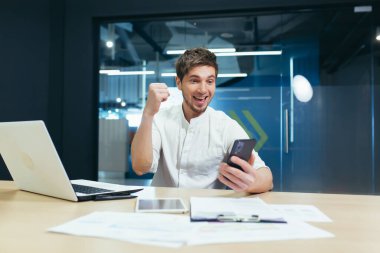 Ofisinde, masasında oturan başarılı genç adam. Elinde bir cep telefonu tutuyor, çevrimiçi piyangoyu kazandığına seviniyor, iyi haberler alıyor. Elleriyle neşe veriyor..