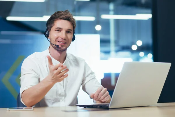 Video conference. A young handsome man in headphones sits in the office behind a laptop and communicates through a video call. He looks at the camera, smiles