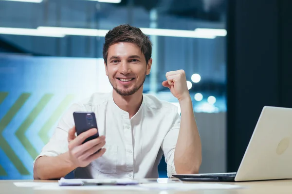 Young coassive successful man sitting in the office at his desk. Holds a mobile phone in his hand, rejoices in winning the online lottery, received good news messages. He shows joy with his hand.