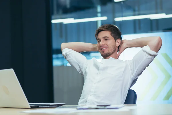 Young handsome man freelancer, businessman, manager in the office at the table resting, doing work during a break, throwing his hands behind his head, smiling