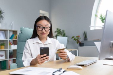 Güzel ve mutlu Asyalı iş kadını ofiste çalışıyor, online alışveriş yapıyor, banka kartı ve telefon tutuyor..