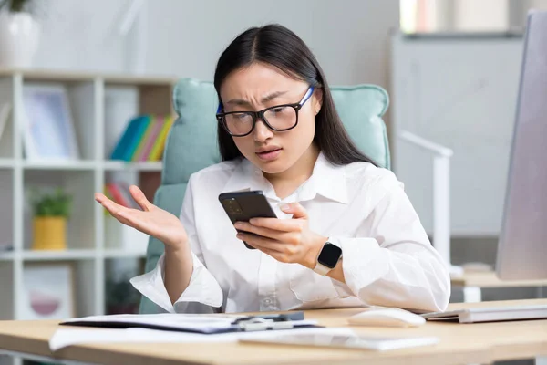 Disappointed and sad asian business woman reading bad news from phone, employee working in office.