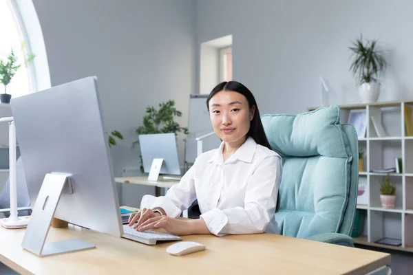 Beautiful Asian woman works at the computer in the office, looks at the camera and smiles, business woman in a white shirt typing on the keyboard.