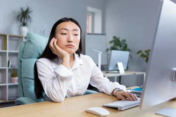 A sad business woman works in a modern office, an Asian woman thinks about the results of work.