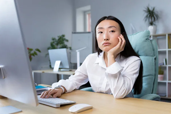 A sad business woman works in a modern office, an Asian woman thinks about the results of work.