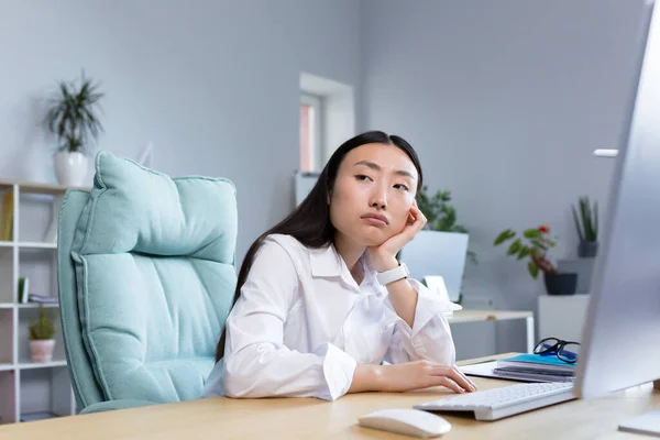 A sad business woman works in a modern office, an Asian woman thinks about the results of work.