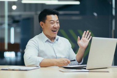 Mutlu ve başarılı Asyalı patron iş arkadaşlarıyla görüntülü konuşma, dizüstü bilgisayar kullanma, kameraya el sallama, iş yerinde iş adamı.