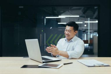 Mutlu ve başarılı Asyalı patron iş arkadaşlarıyla görüntülü konuşma, dizüstü bilgisayar kullanma, kameraya el sallama, iş yerinde iş adamı.