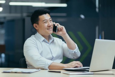 Bilgisayarlı, iş adamı gülümseyen ve telefonda konuşan başarılı bir Asyalı girişimci..