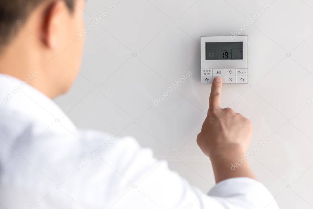 Asian businessman in office heat, chooses cooling mode of air conditioner on control panel on white wall