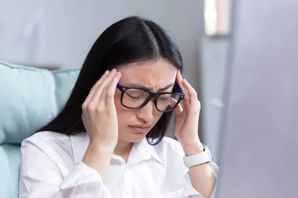 Exhaustion at work. A young Asian woman in glasses holds her head, feels pain, grimaces. Sitting in the office at the desk, working at the computer, exhausted. Businesswoman, accountant, freelancer.