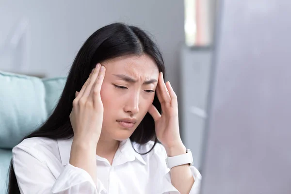 Close up photo. Exhaustion at work. A young Asian woman holds her head, feels pain, grimaces. Sitting in the office at the desk, working at the computer, exhausted. Businesswoman, accountant