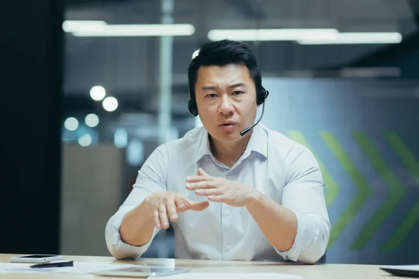Serious and disappointed asian businessman with headset for video call, looking at web camera, unhappy talking online meeting