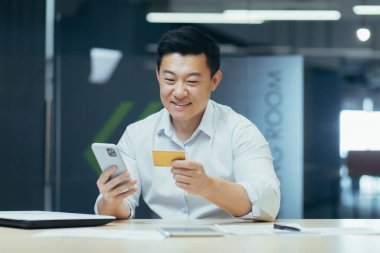 Ofisteki başarılı Asyalı işadamı online alışveriş yapıyor. Adamın elinde telefon ve banka kartı var.