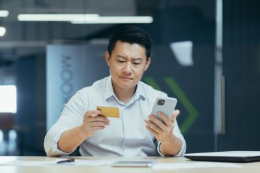 Hüsrana uğramış Asyalı işadamı kredi kartı ve telefonuyla ofiste çalışıyor, online alışveriş yapmaya çalışıyor ve banka havalesi yapıyor.