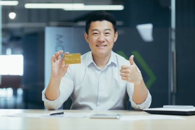 Ofiste çalışan Asyalı işadamı, elinde kredi kartı olan bankacı kameraya bakıyor ve gülümsüyor.