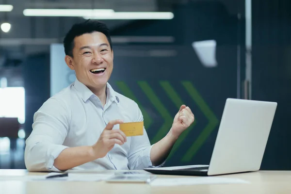 Happy Asian businessman smiling and looking at camera, holding bank credit card for purchase in online store, celebrating successful banking transaction, man in office at work