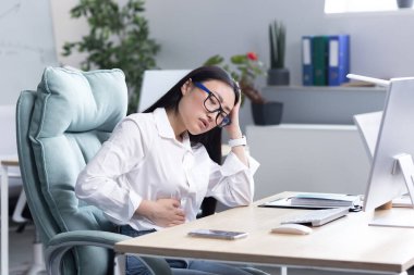 İş yerinde karın ağrısı var. Gözlüklü, genç ve güzel bir Asyalı kadın bilgisayarın ofisinde çalışıyor, karnında ağrı hissediyor, elini karnına dayayıp, acı içinde kıvranıyor..