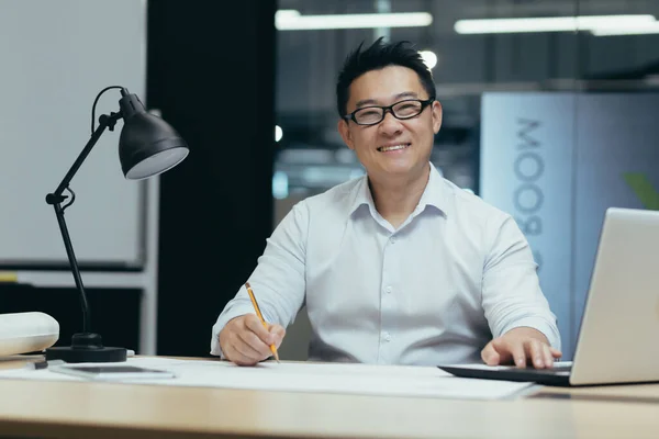 Portrait of Asian designer, creative work, man looking at camera and smiling, working in architecture office on plan drawing