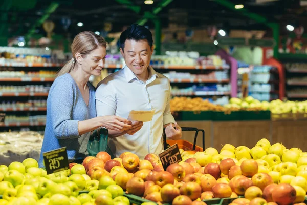 happy mixed race asian couple family man and woman choosing fruits or vegetables in grocery store supermarket. Joint daily shopping together. Buyers customer select product pick an apple in eco bag