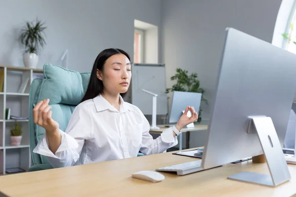 Break and meditation at work. Young beautiful asian businesswoman wearing glasses sitting at desk in office with eyes closed, holding hands in lotus yoga pose, resting, relaxed.