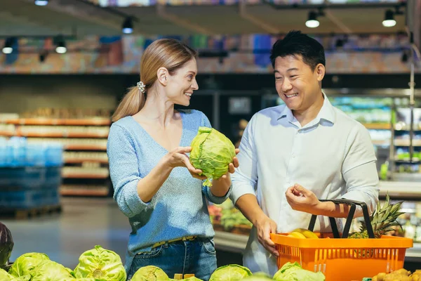 Happy mixed race asian couple family man and woman choosing vegetables in grocery store supermarket. make a purchase daily shopping. Customer select product pick an cabbage in eco bag in food market