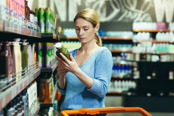 Female chooses and examines product on the supermarket shelves. household chemicals or body care goods detergents in store. Woman customer background reads and inspects a label shampoo make a purchase