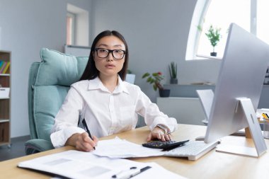 Work with documents. Portrait of a young beautiful business woman Asian accountant works with documents and reports. Sitting at the desk in the office, writing.He looks at the camera.