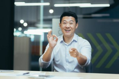 Çevrimiçi webinar. Aktif bir Asyalı genç iş adamının portresi. Kamerada internetten bir web sitesi yönetiyor, modern bir ofiste bir masaya oturuyor, konuşuyor, anlatıyor, elleriyle işaret ediyor, gülümsüyor..