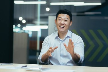 Çevrimiçi webinar. Aktif bir Asyalı genç iş adamının portresi. Kamerada internetten bir web sitesi yönetiyor, modern bir ofiste bir masaya oturuyor, konuşuyor, anlatıyor, elleriyle işaret ediyor, gülümsüyor..