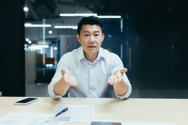 Çevrimiçi iş eğitimi. Aktif bir Asyalı genç iş adamının portresi. Kamerada online iş eğitimi veriyor, modern bir ofiste masaya oturuyor, konuşuyor, açıklıyor, ellerini sallıyor, gülümsüyor.